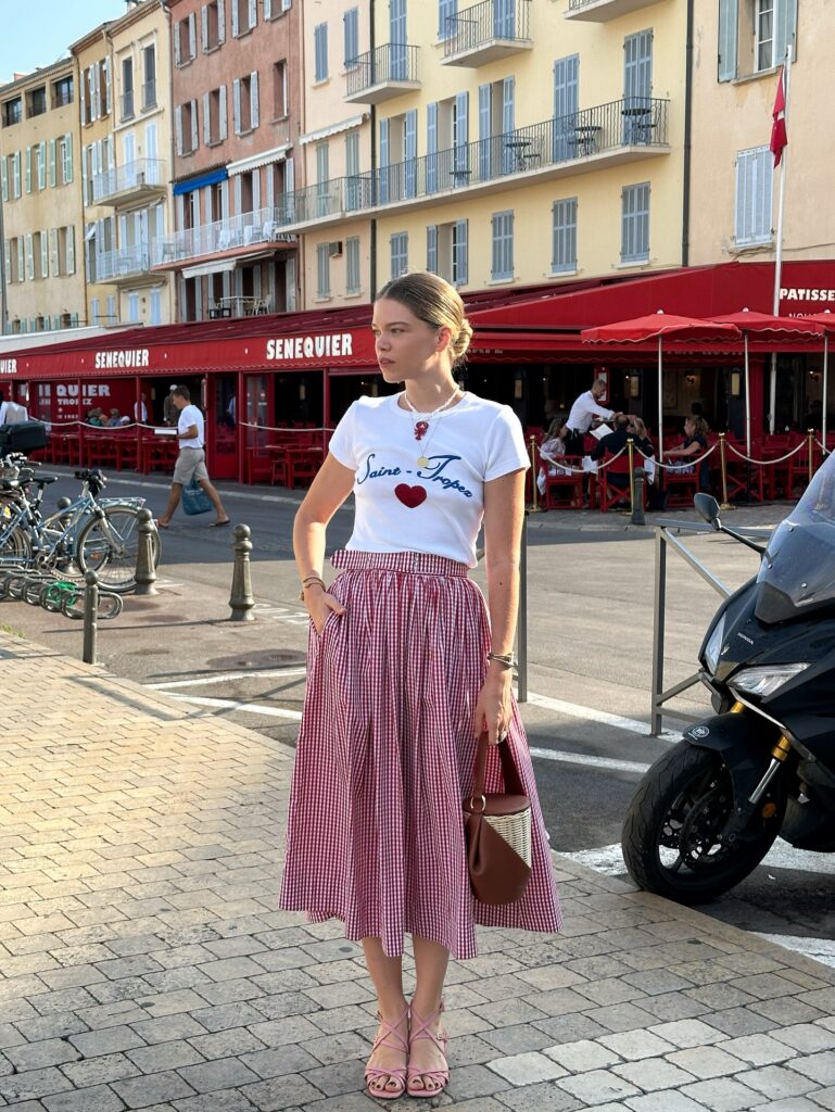 Woman in casual fashion with a heart-printed tee and checkered skirt, standing on a lively street in a coastal town.