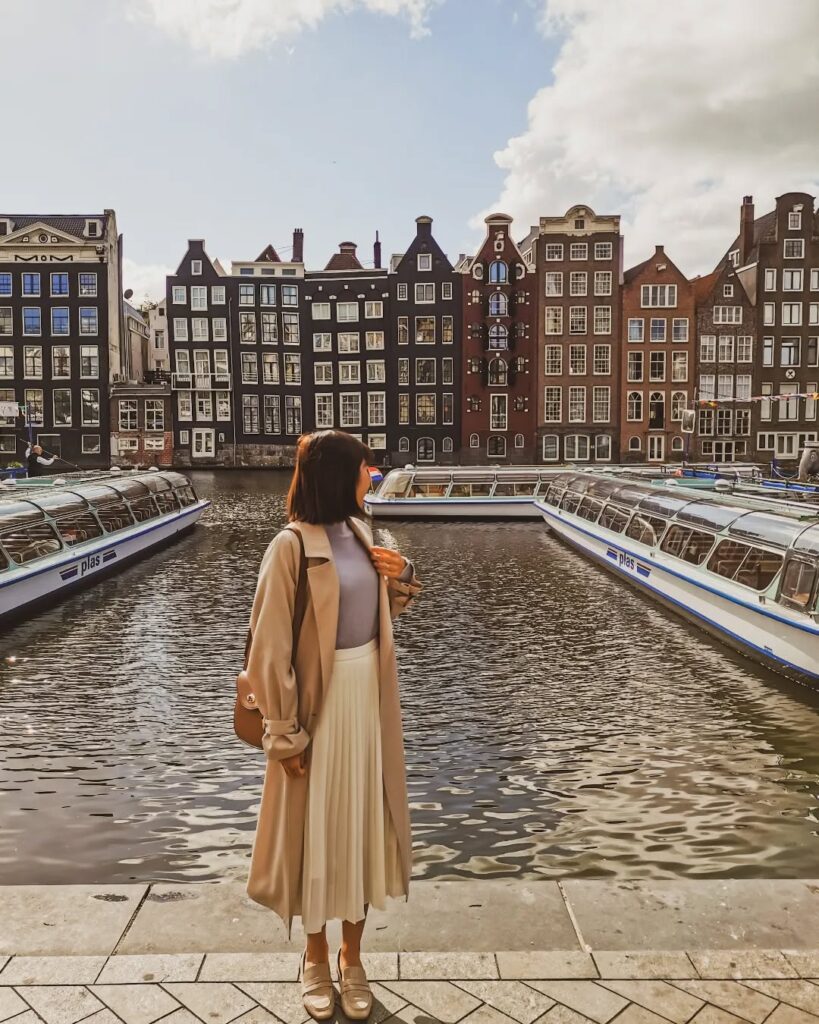 Woman in a beige coat stands by the water in Amsterdam, with charming canal houses and boats in the background.