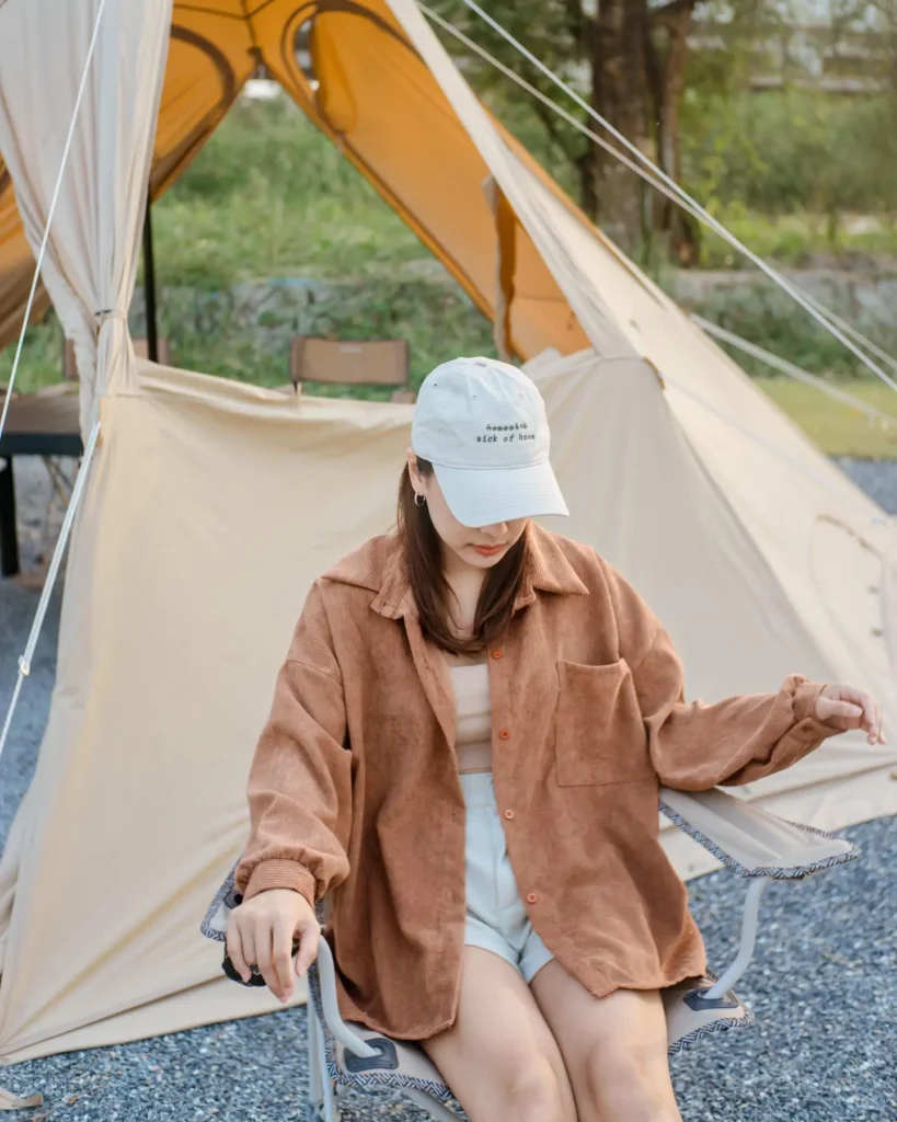 Woman in a cozy oversized shirt and cap, relaxing by a tent in a serene outdoor setting. Perfect for camping style inspiration.