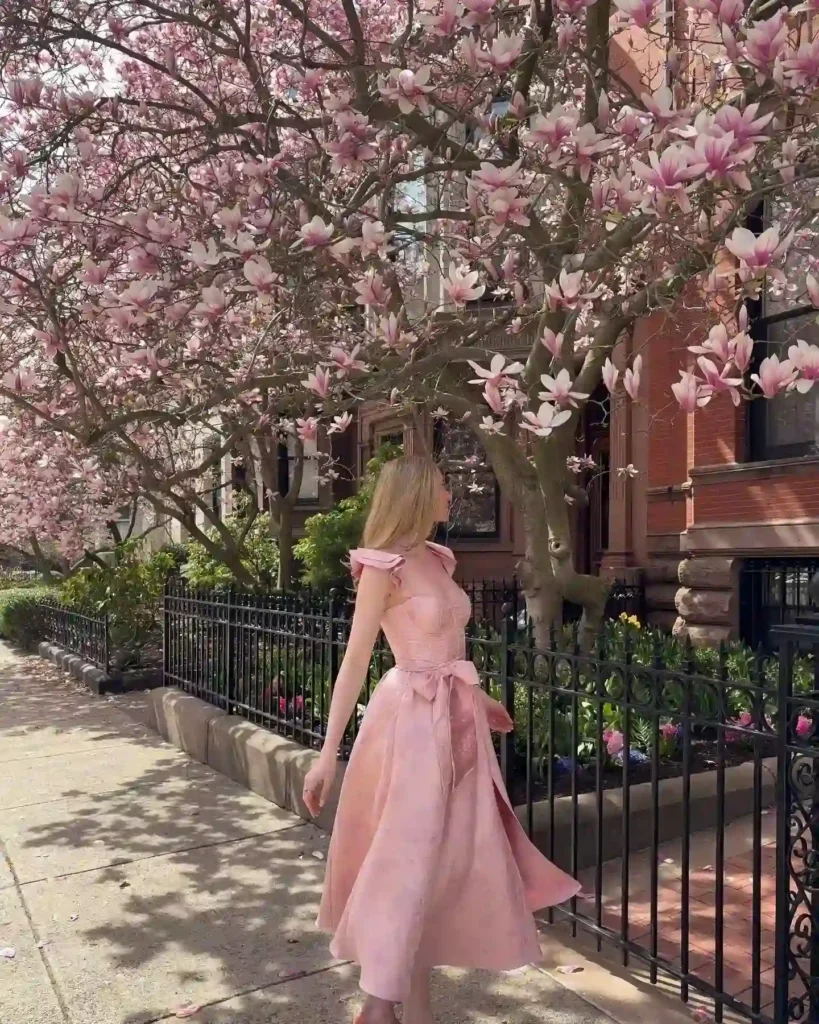 Woman in a pink dress walking past blooming magnolia trees on a sunny urban sidewalk. Spring fashion inspiration.