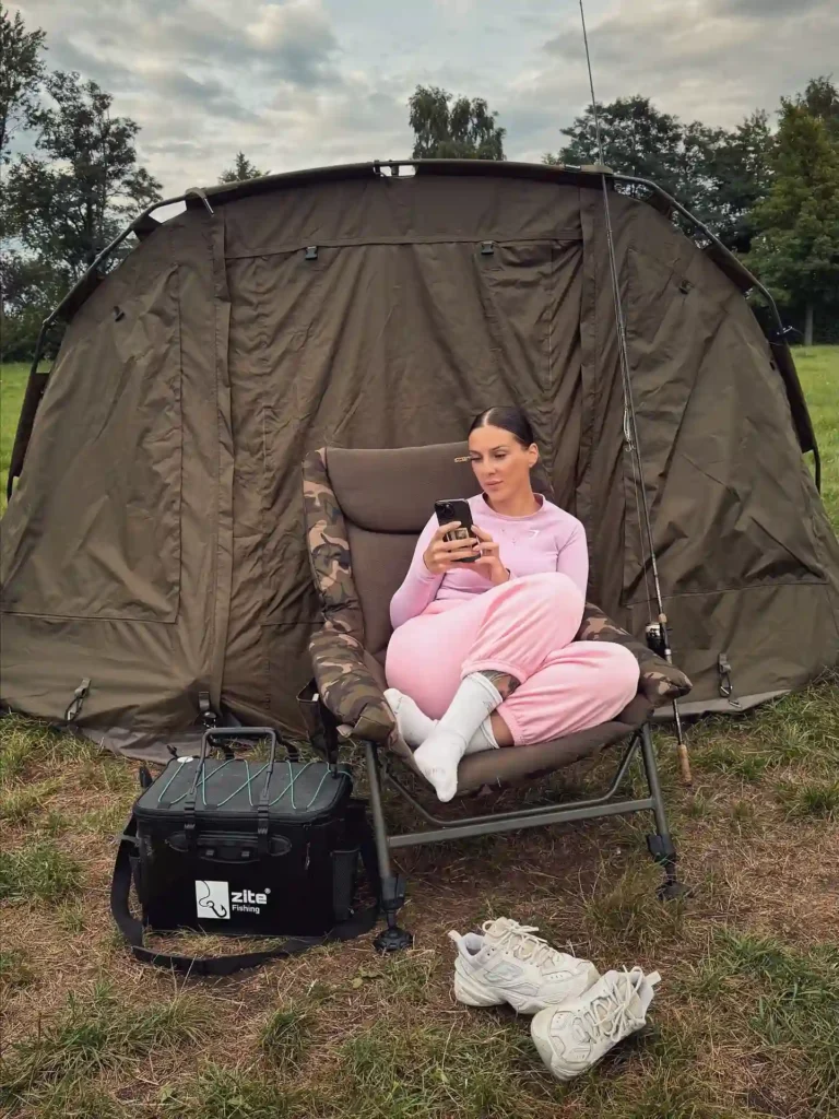 Young woman in pink loungewear sitting on a camping chair, using a smartphone outside a fishing tent.
