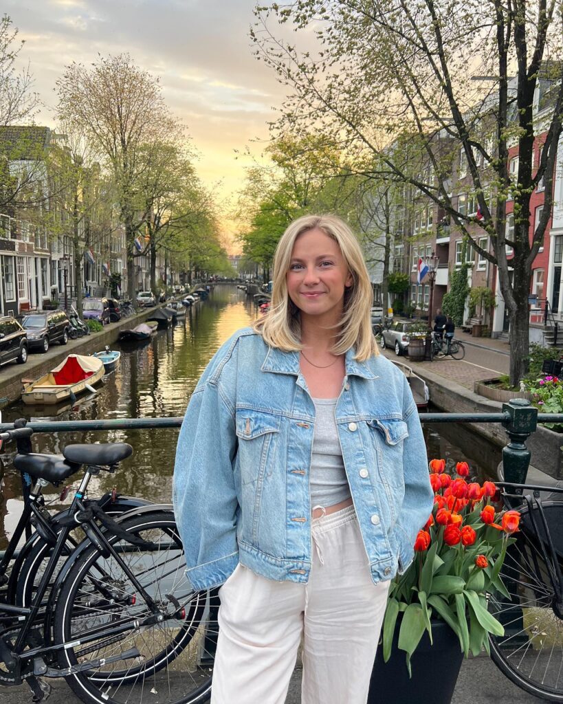 Young woman in a denim jacket poses by a canal with blooming tulips and bicycles in a picturesque cityscape.