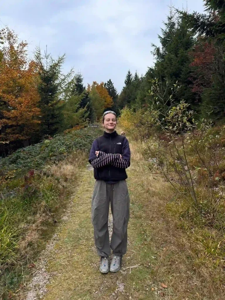 A person standing confidently on a forest path, surrounded by trees and autumn foliage, enjoying nature.
