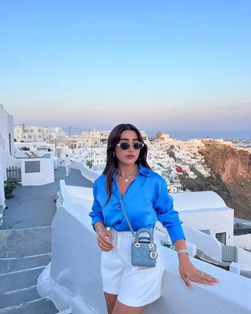 A fashionable woman in a blue shirt and white shorts poses against a stunning Santorini backdrop.