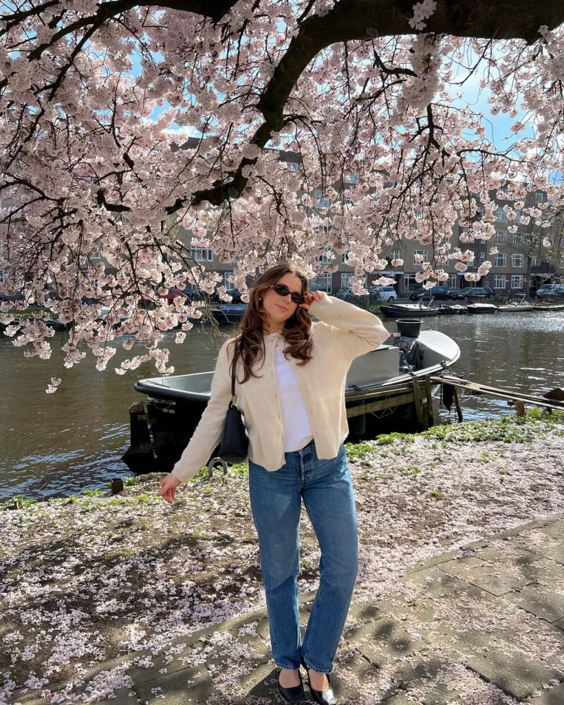 Young woman in sunglasses posing under cherry blossoms by a canal, wearing a cream cardigan and blue jeans. Springtime vibes.