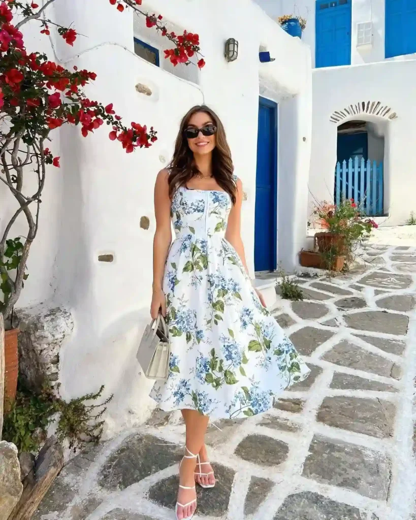 Woman in a floral dress and sunglasses smiles on a picturesque street with white walls and blue doors.