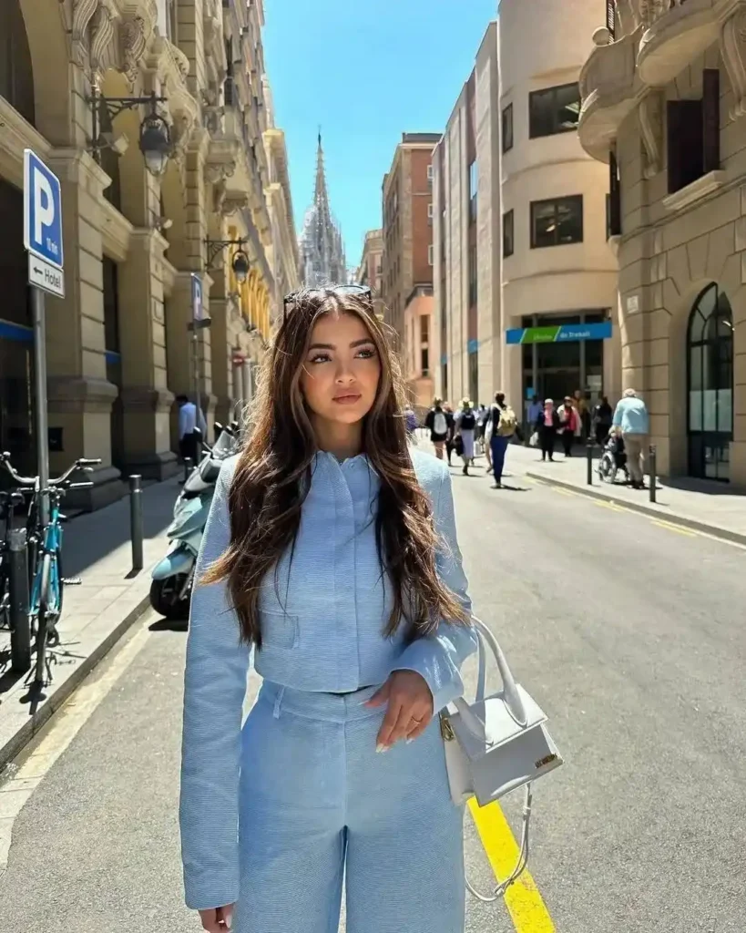 Young woman in a chic blue suit stands on a sunny street with historic buildings, showcasing urban fashion and style.