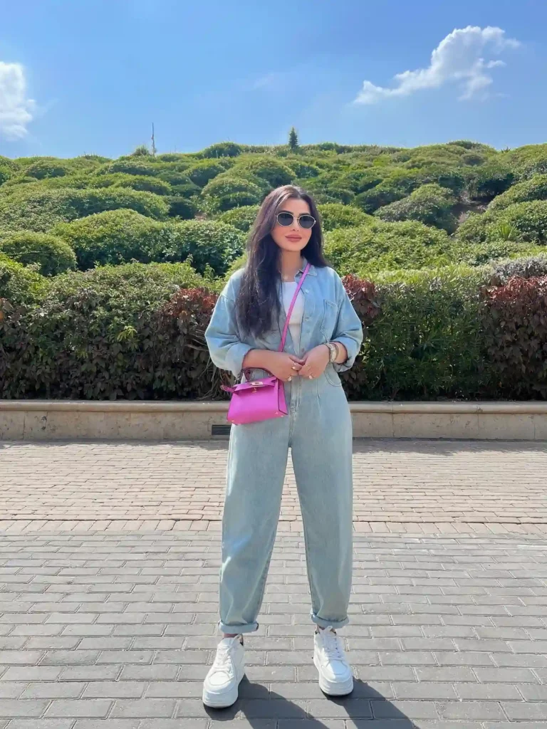 Person standing outdoors in a light denim outfit with sunglasses, pink handbag, and white sneakers against a green backdrop.