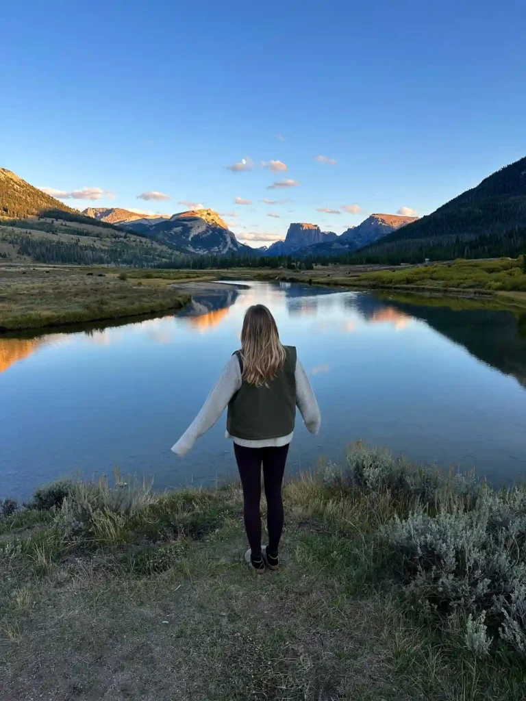 Person standing by a serene lake surrounded by mountains, enjoying a peaceful sunset view in nature.