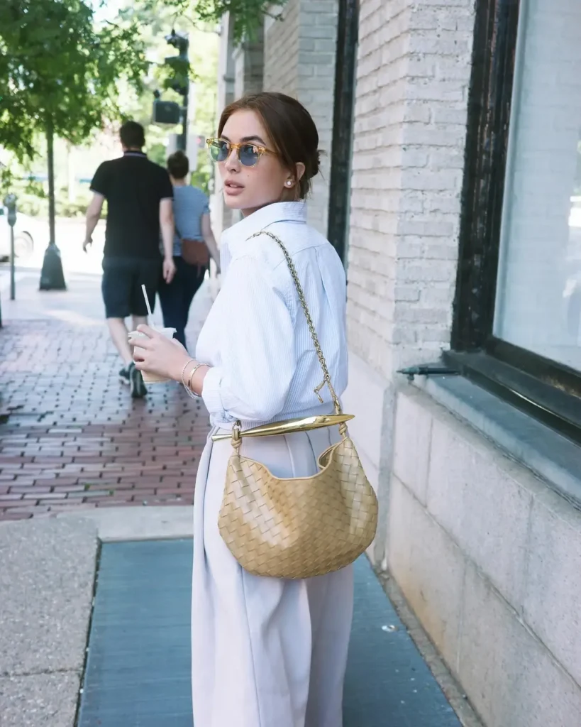 Stylish woman in sunglasses and a white shirt, holding a drink while walking down a city street with a chic handbag.