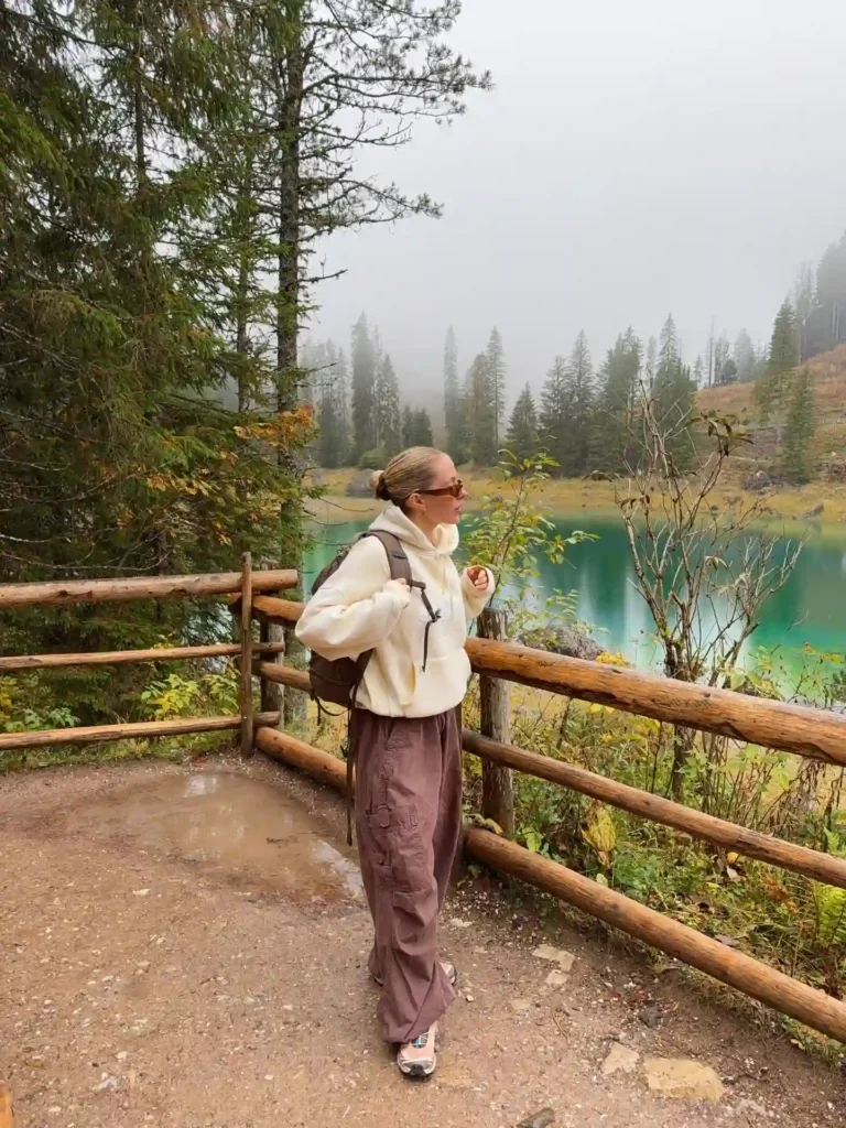 Woman in casual attire stands by a scenic lake surrounded by misty trees, enjoying a peaceful outdoor moment.