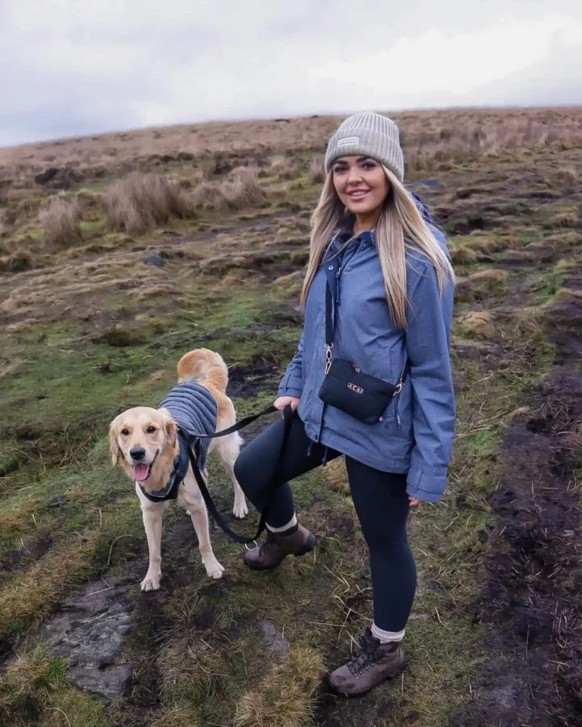 Young woman in a blue jacket and hat walking her happy dog on a scenic trail, surrounded by grassy hills.