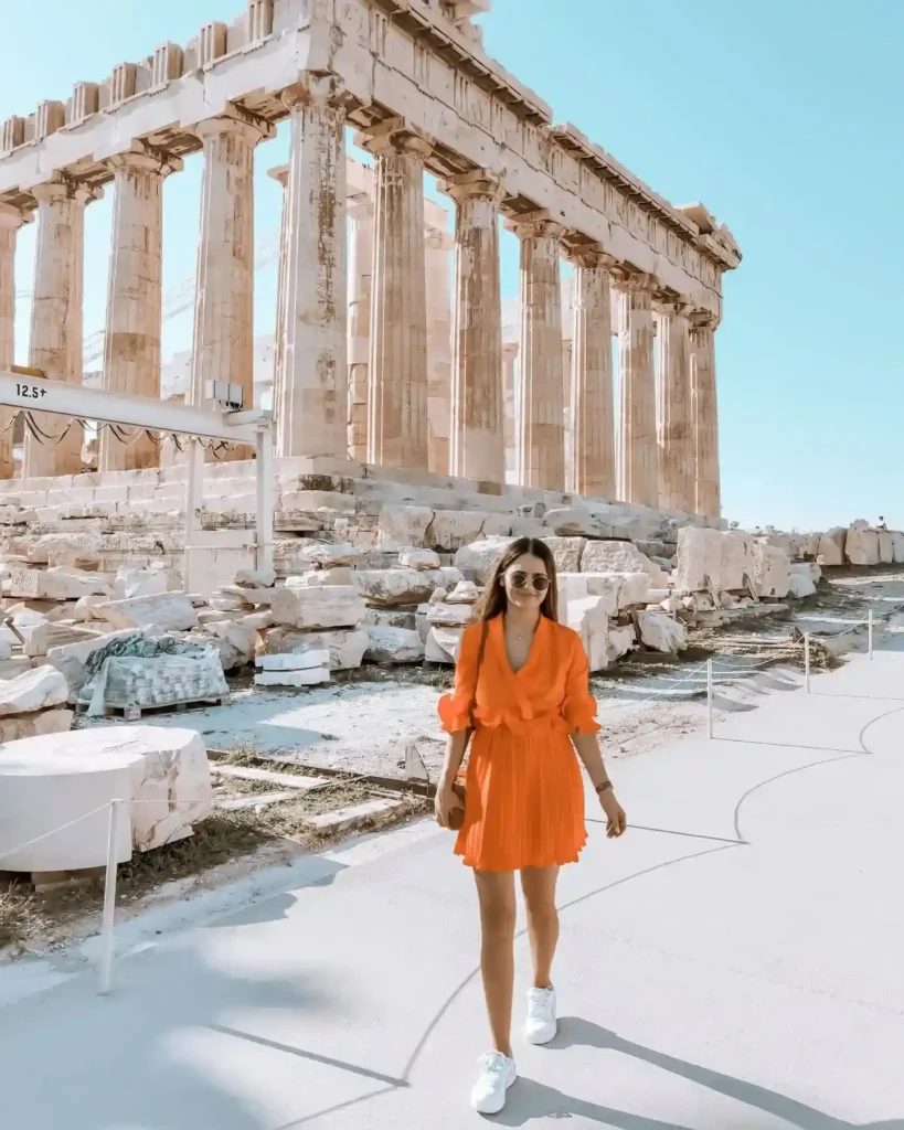 Tourist in an orange dress walking by the Parthenon, Athens, with ancient ruins and a clear blue sky in the background.