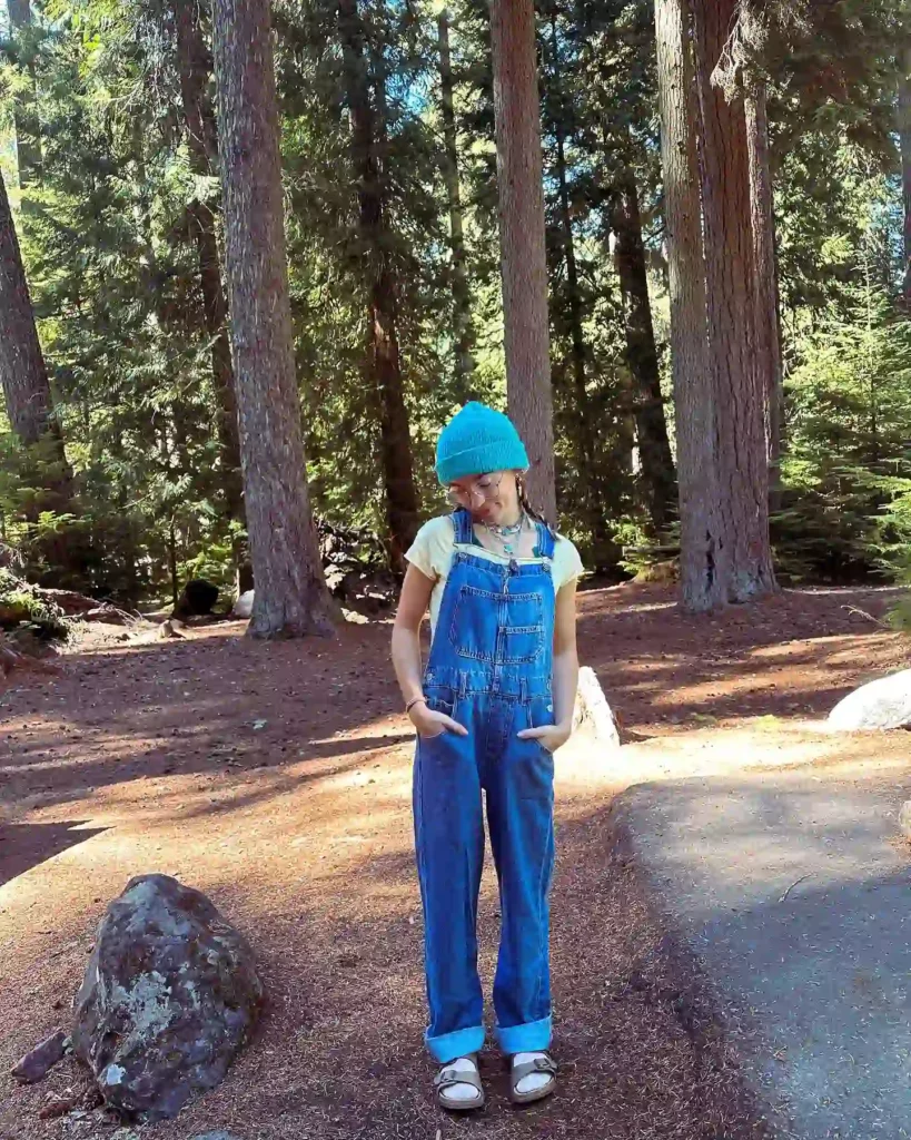 Child in blue overalls and a blue hat standing in a serene forest with tall trees, enjoying a sunny outdoor day.