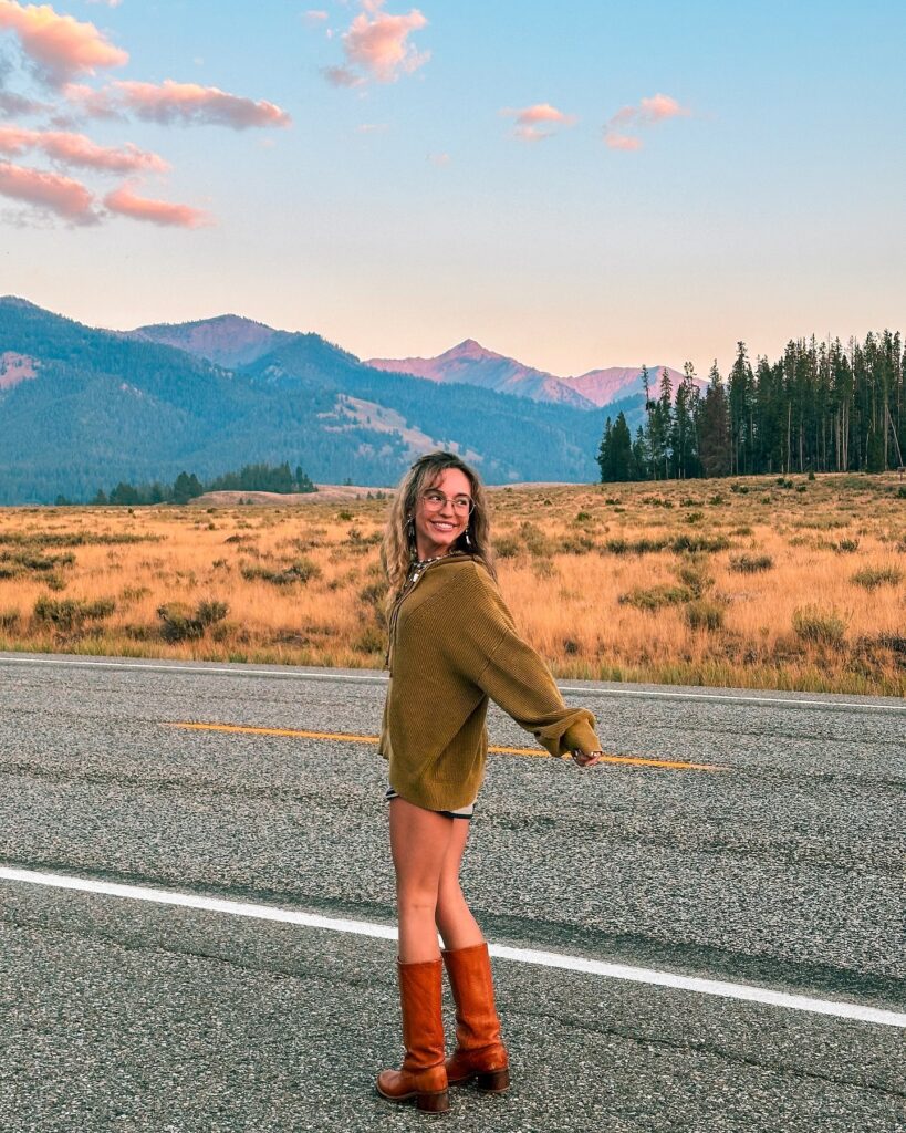 Young woman in a cozy sweater and brown boots poses on a scenic road against a backdrop of mountains and blue sky.
