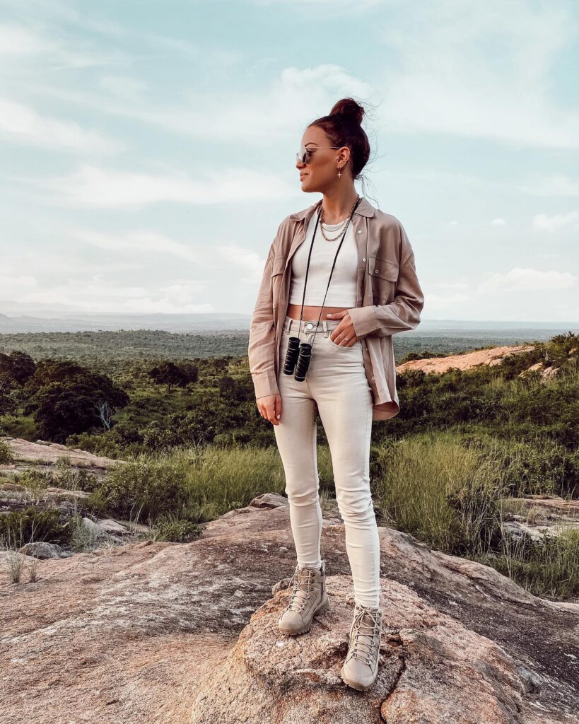 Young woman in stylish outdoor attire stands on a rock, enjoying a scenic view with binoculars around her neck.