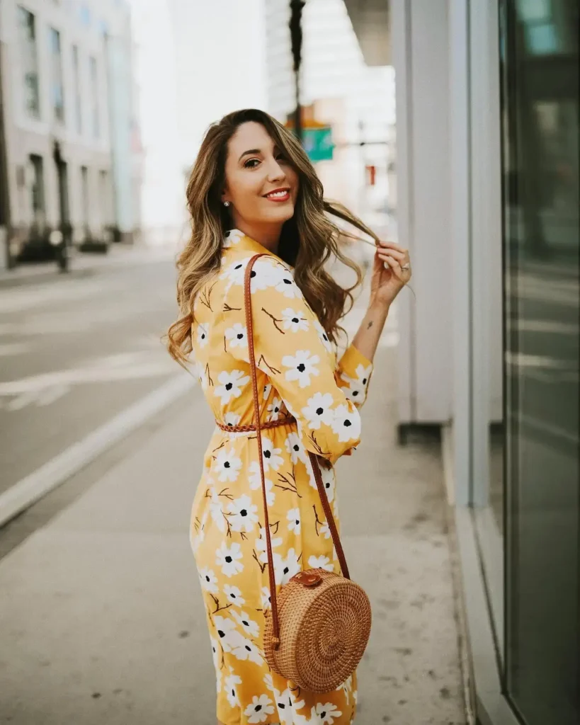 A woman in a yellow floral dress smiles and walks down a city street, holding a round straw bag.