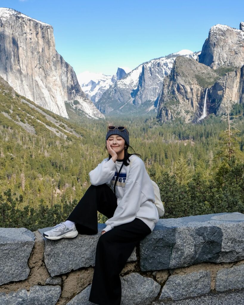 Young woman in a gray sweatshirt poses on a stone wall with a scenic mountain backdrop, featuring snow-capped peaks and greenery.