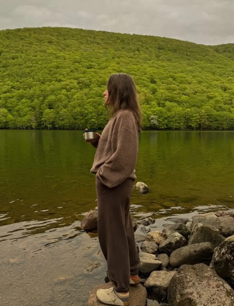 Person standing by a serene lake, surrounded by lush green hills, sipping from a metal mug on a cloudy day.