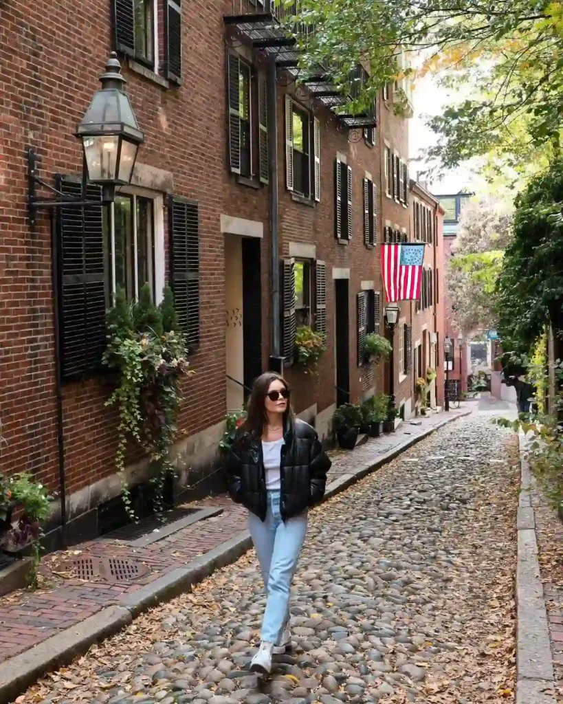 Woman walking down a cobblestone street lined with historic brownstone buildings and an American flag. Autumn setting.
