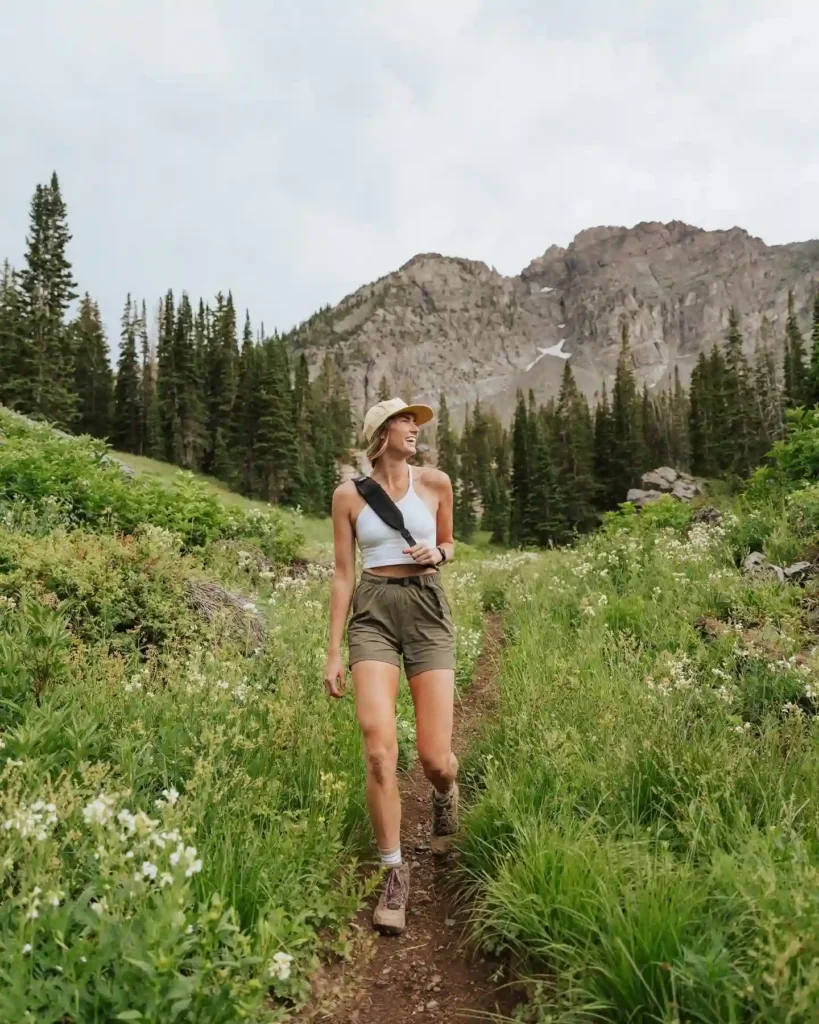 Woman hiking on a trail surrounded by lush greenery and mountains under a cloudy sky, enjoying nature's beauty.