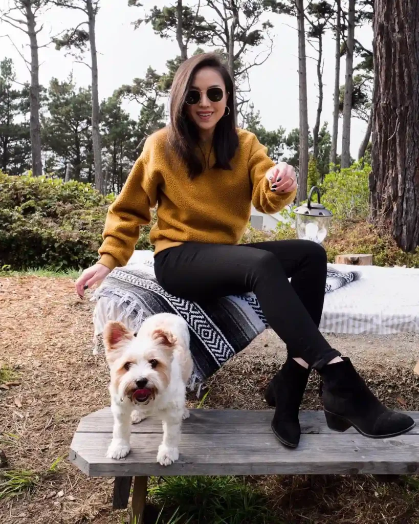 Woman in stylish outfit sits on a bench outdoors with a cute dog, surrounded by trees and a cozy blanket.