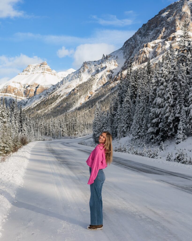 Woman in a pink sweater and jeans poses on a snowy mountain road with trees and stunning peaks in the background.