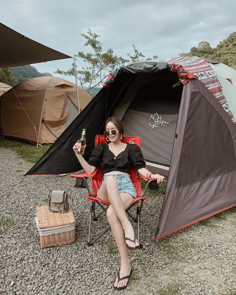 Young woman relaxing in a camping chair with a drink, surrounded by tents and nature, enjoying a serene outdoor getaway.