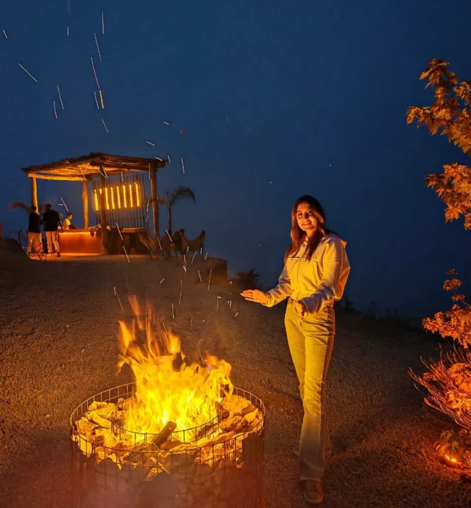 A woman smiles by a cozy campfire at dusk, with a warm glow from a nearby structure in a serene outdoor setting.