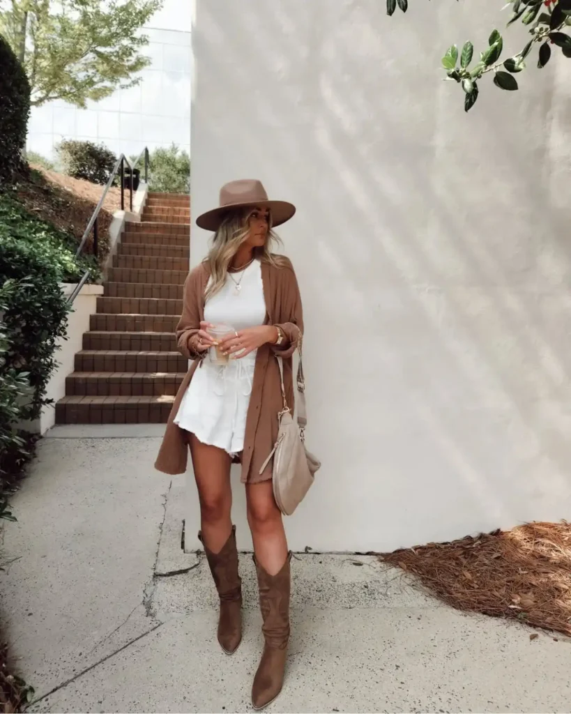 Stylish woman in a brown hat, cozy cardigan, and trendy cowboy boots standing near stairs and greenery.