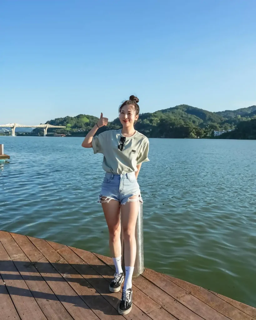 Young woman in a casual outfit stands by the water, smiling and giving a thumbs up on a sunny day.