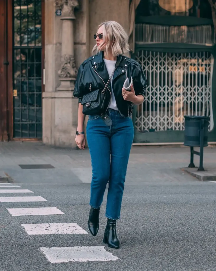Fashionably dressed woman in a leather jacket and jeans walking confidently across a street. Stylish urban backdrop.