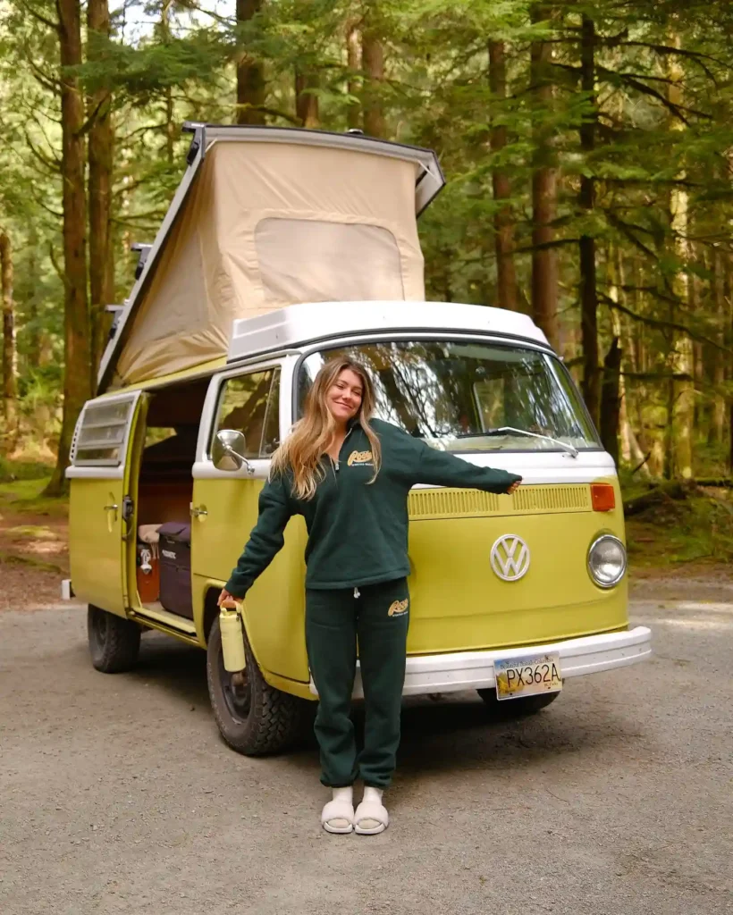 Young woman in a green outfit poses by a vintage VW camper van in a lush forest setting, promoting outdoor adventures.