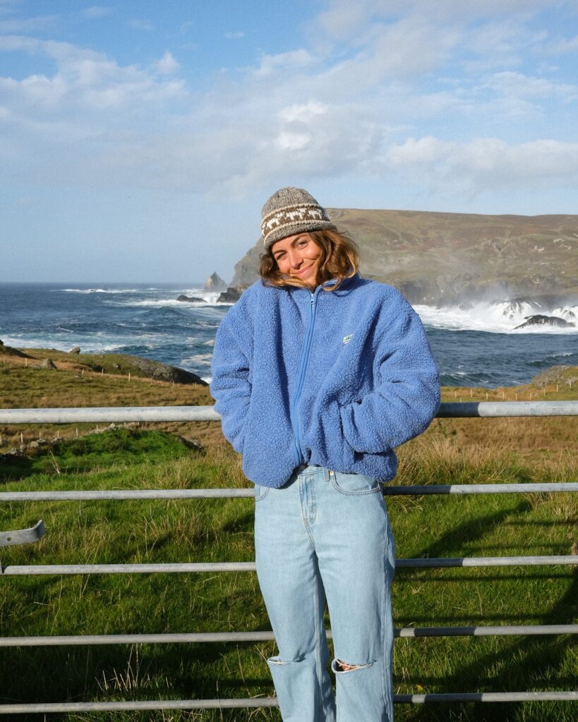 Smiling woman in a blue fleece jacket and knit hat stands by the ocean, enjoying a sunny day with scenic cliffs behind her.