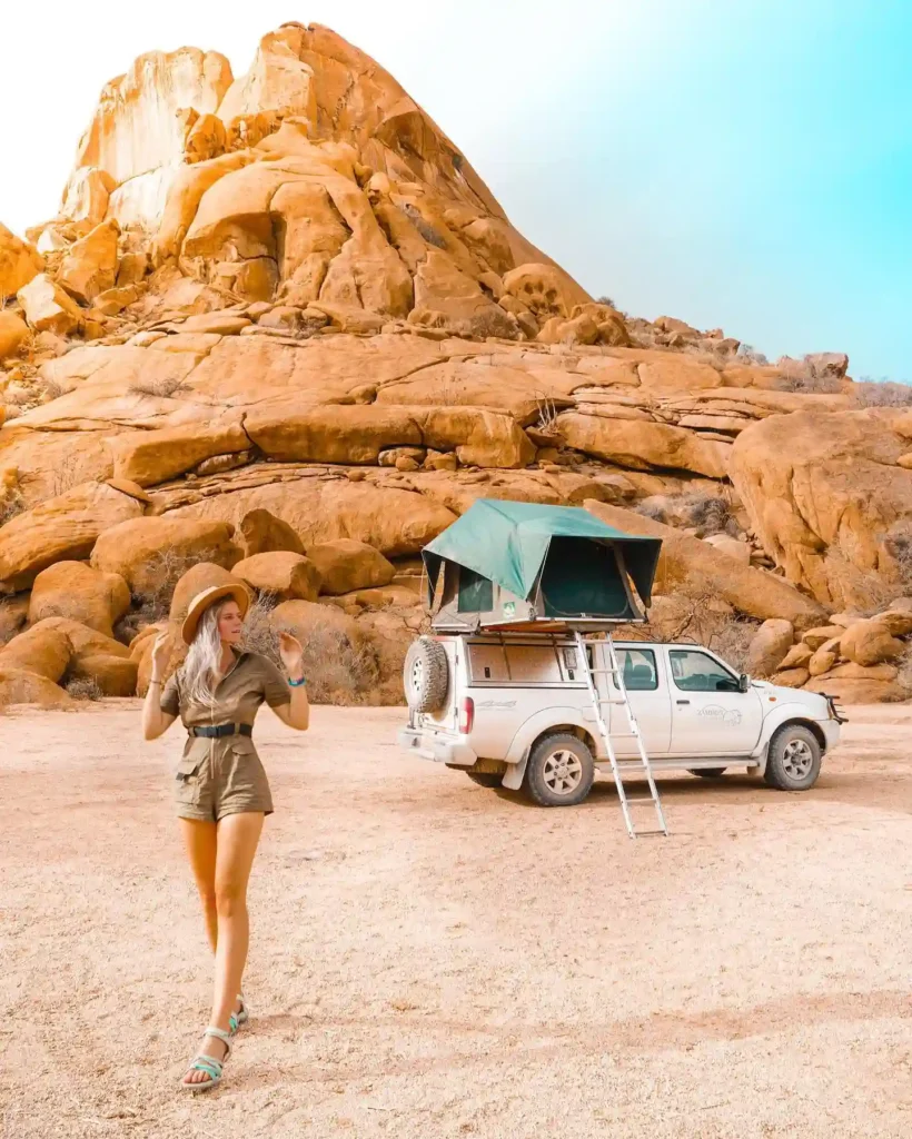 Woman in a hat standing near a truck with a rooftop tent against a rocky landscape, enjoying an outdoor adventure.