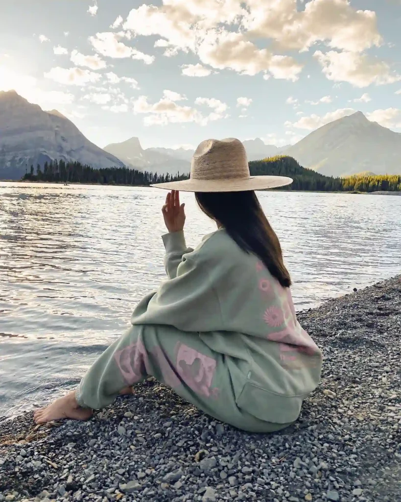 Woman in oversized sweater and hat relaxing by a serene lake surrounded by mountains, enjoying nature at sunset.