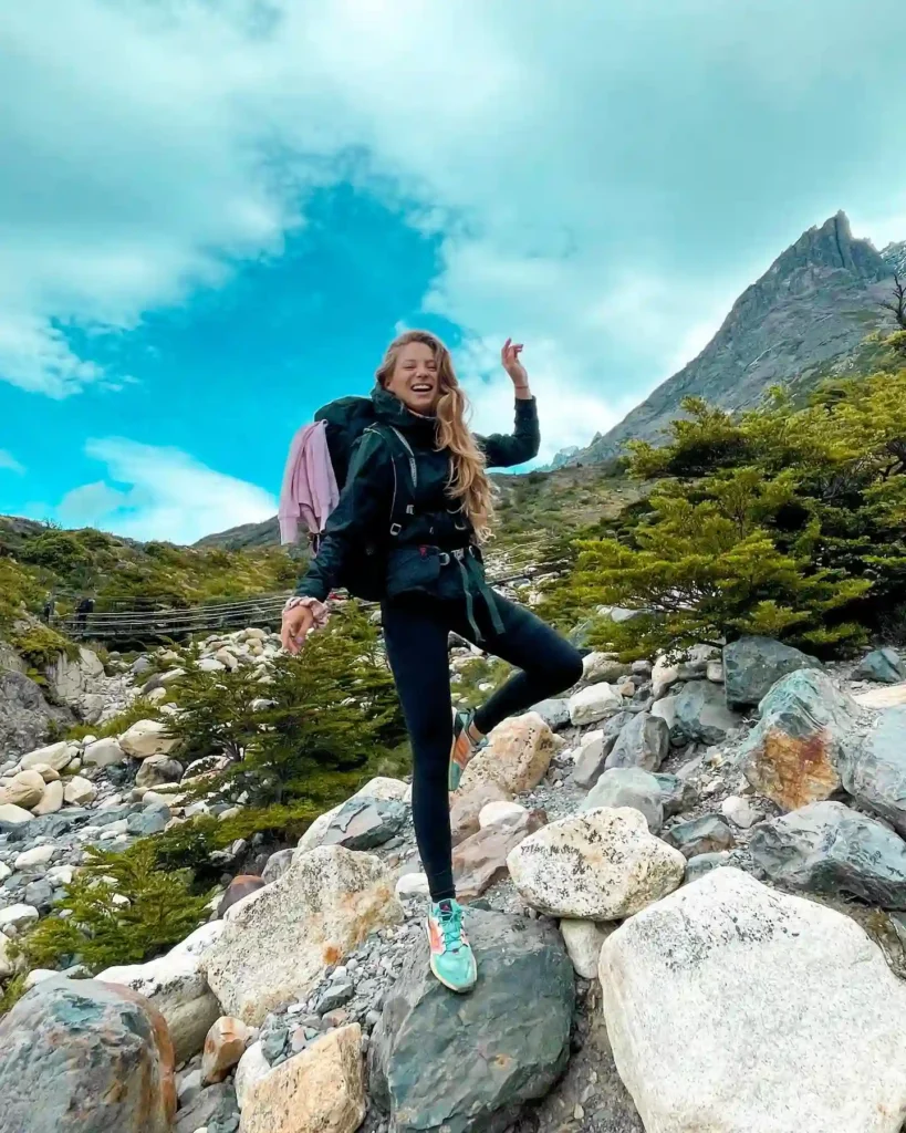 A happy woman with long hair joyfully hiking over rocky terrain with mountains and blue skies in the background.