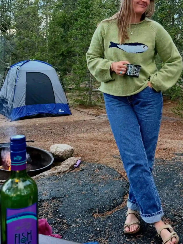 Woman in a cozy sweater stands near a campfire, bottle of wine, and a tent in a picturesque forest campsite.