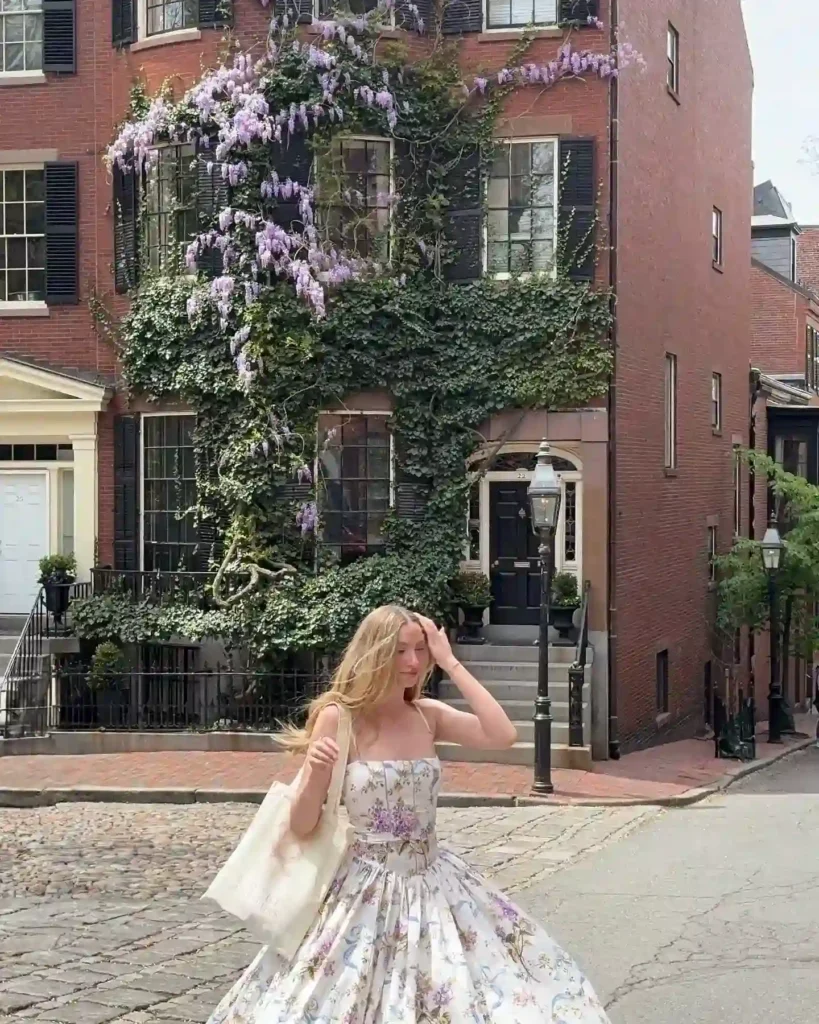 A woman in a floral dress walks past a charming brick house adorned with purple flowers and lush greenery.
