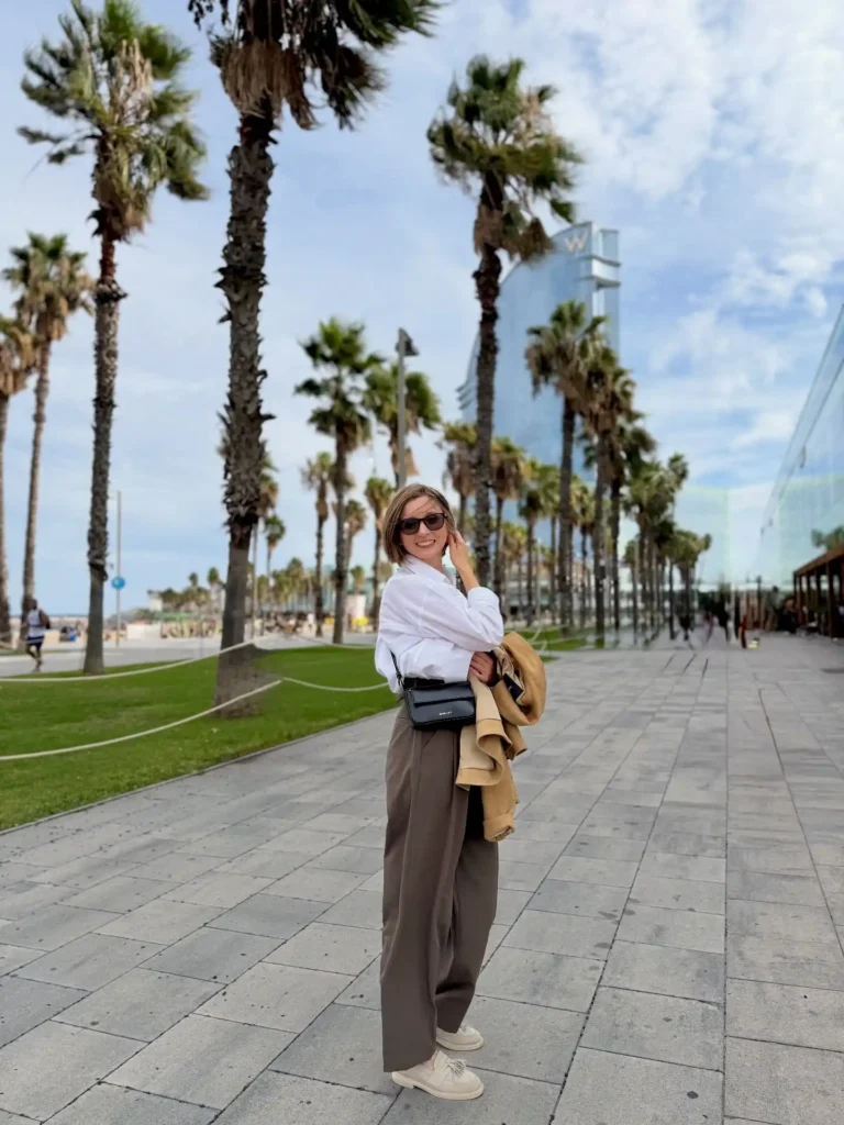 Woman in casual outfit posing on a palm-lined walkway near the beach, enjoying a sunny day in a modern city.