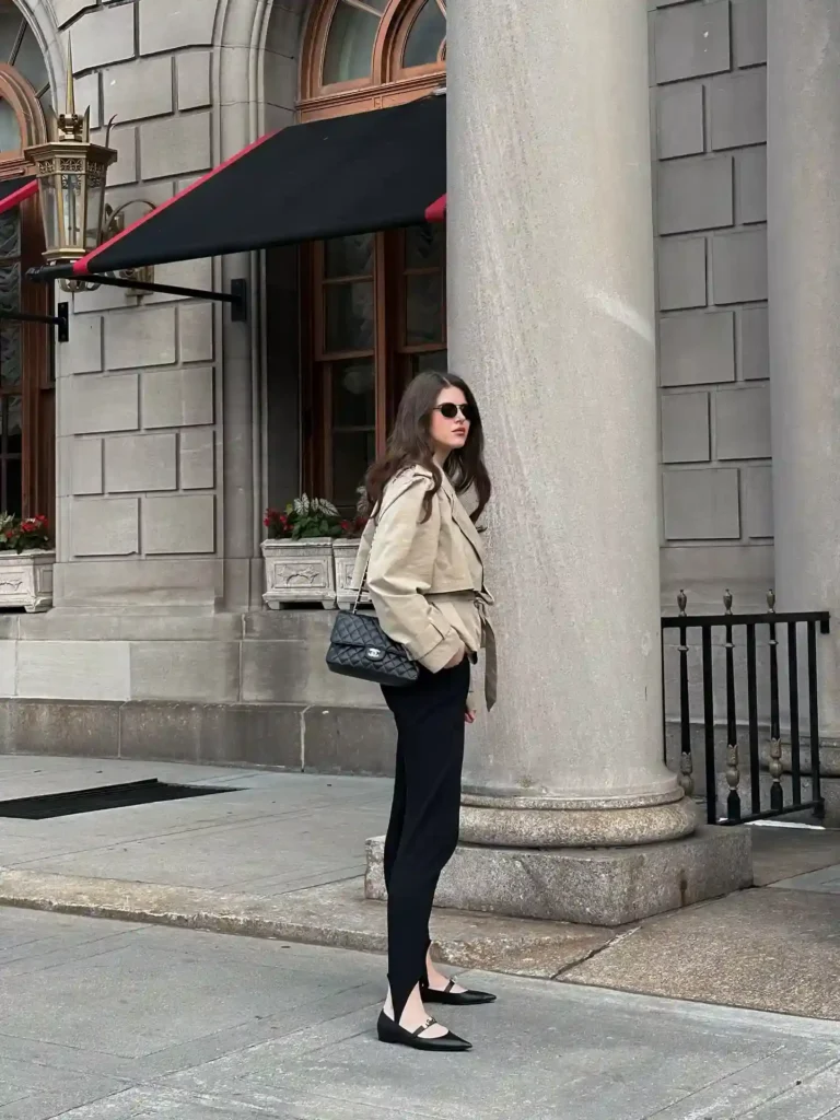 Fashionable woman in a beige trench coat and black pants stands stylishly outside a historic building on a city street.