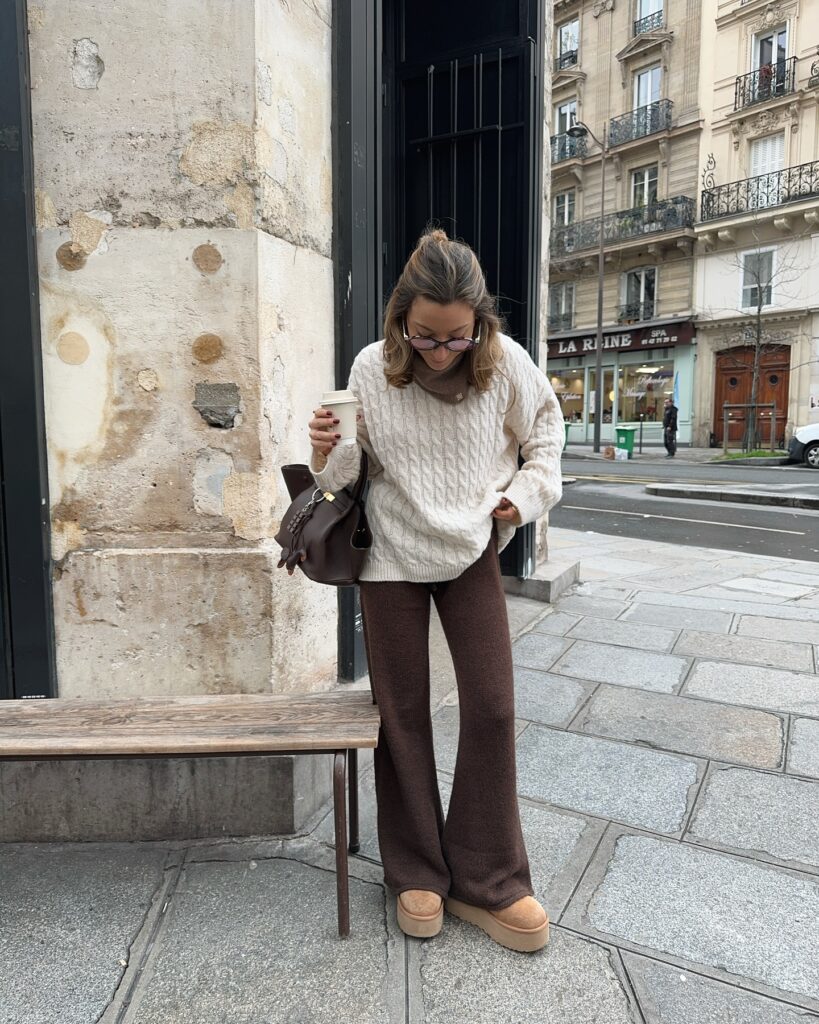 Woman in a cozy cream sweater and brown pants enjoys coffee on a city street near historic buildings.