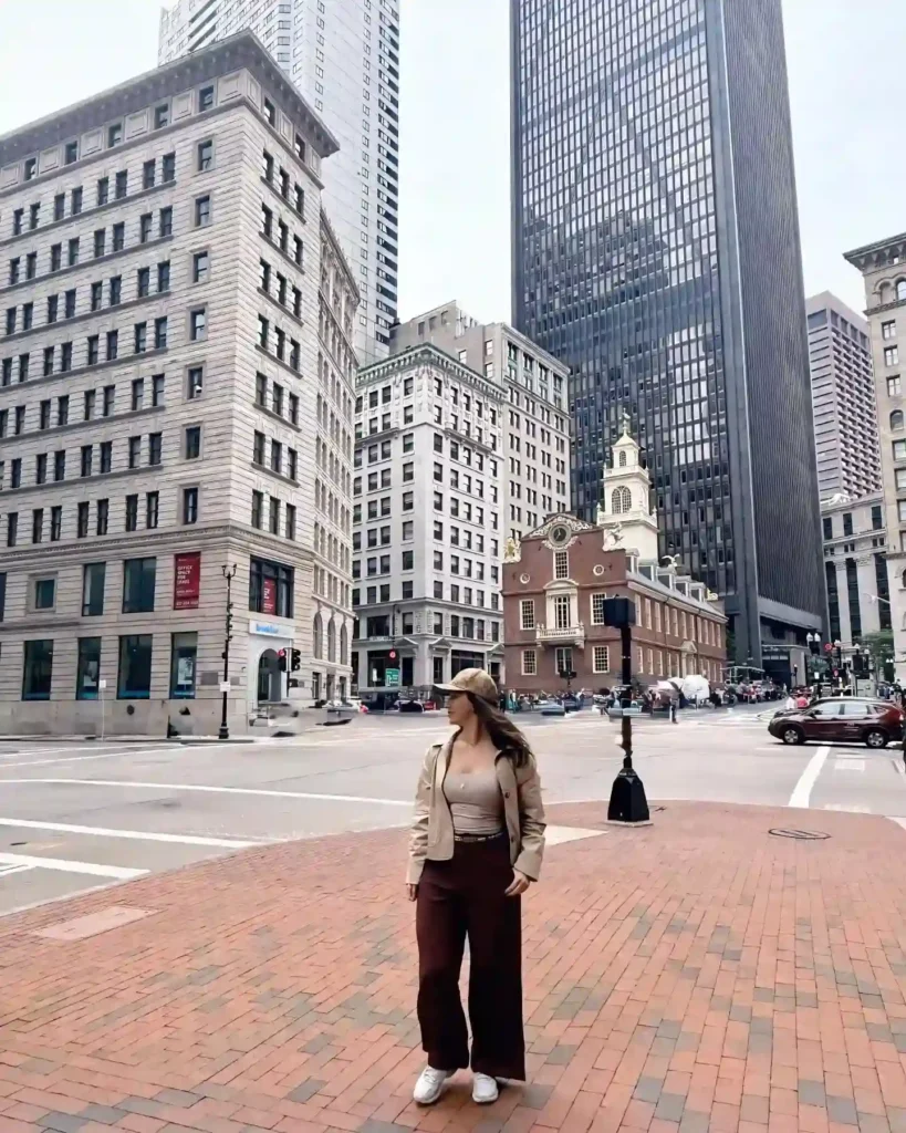 A woman in stylish attire stands on a downtown street, surrounded by historic and modern buildings.