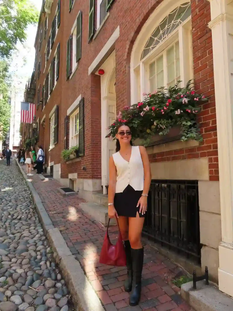 Young woman in a stylish outfit poses on a charming cobblestone street with brick buildings and flowers.