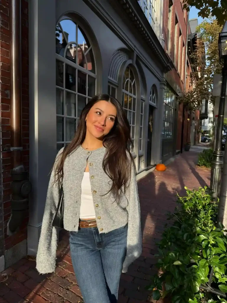 Young woman in a gray cardigan smiling on a sunny street with brick pathways and blooming greenery.