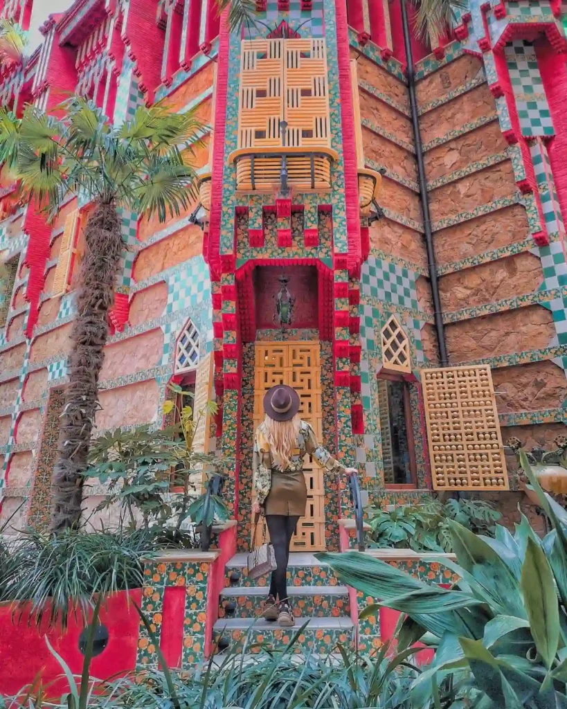 Woman in a stylish outfit walks up to a vibrant, intricately designed building surrounded by lush greenery.