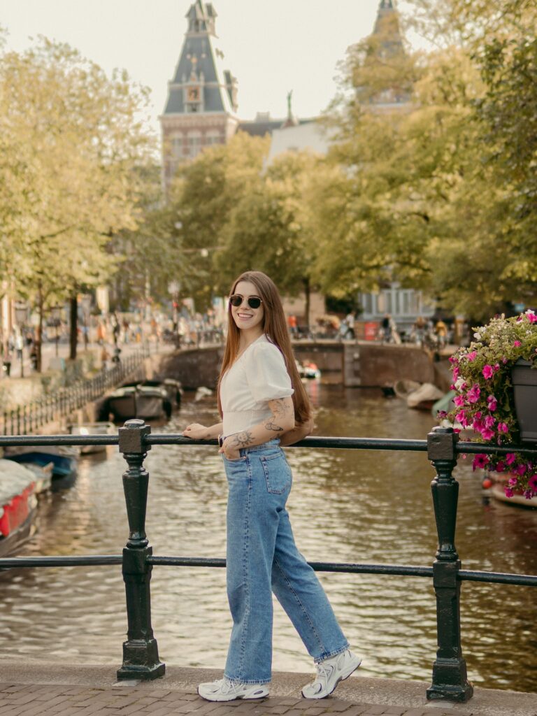 Young woman in stylish attire stands by a canal in Amsterdam, surrounded by greenery and historic architecture.