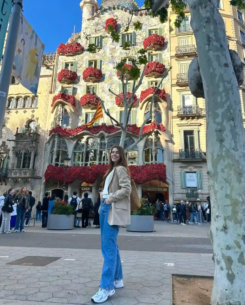 Young woman smiling in front of Casa Batlló, adorned with red flowers, surrounded by visitors in Barcelona.