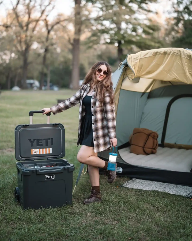 Young woman in a plaid jacket with a YETI cooler and camping tent, enjoying outdoor adventure in nature.