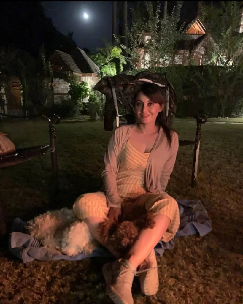 Woman sitting outdoors at night, cuddling two dogs under the moonlight, surrounded by a serene garden setting.